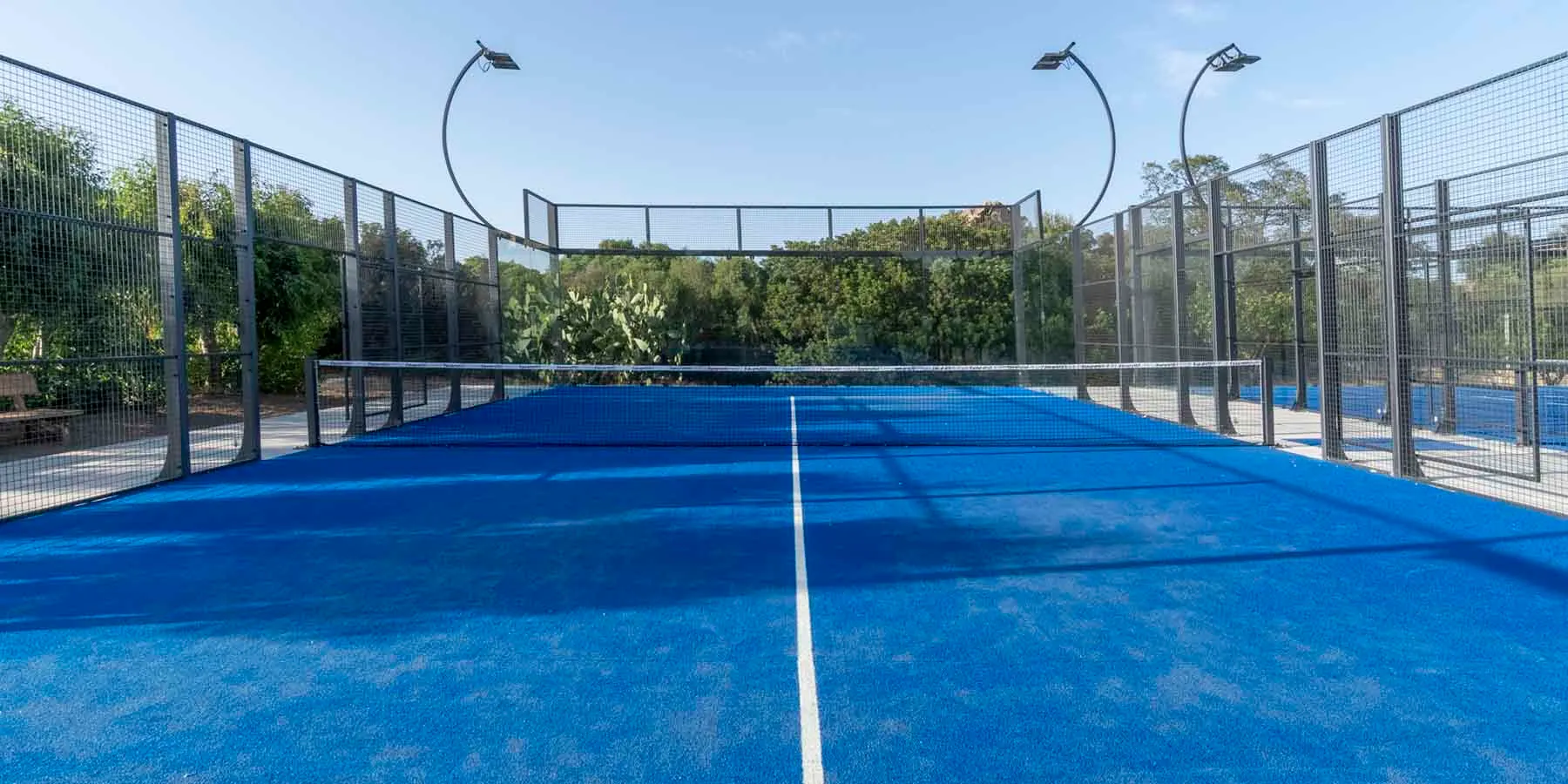 Empty panoramic padel court at Lu Nibareddu, blue turf and glass walls framed by Mediterranean vegetation