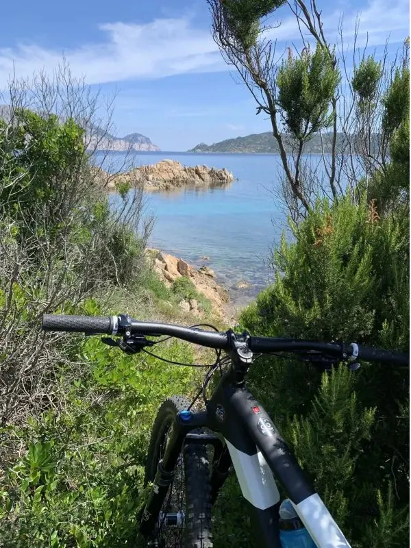 Bike handlebars framing a turquoise cove with pink granite rocks between Porto Taverna and Cala Brandinchi