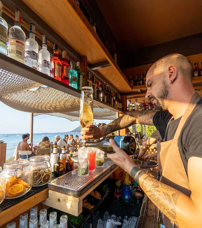Bartender of the Tavernetta beach bar pouring a cocktail, the sea behind the counter