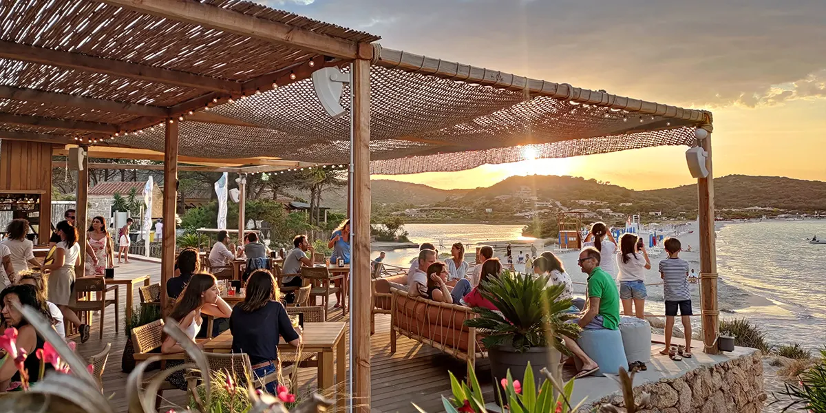 Guests at the Tavernetta Beach bar deck at sunset, with Porto Taverna bay glowing behind
