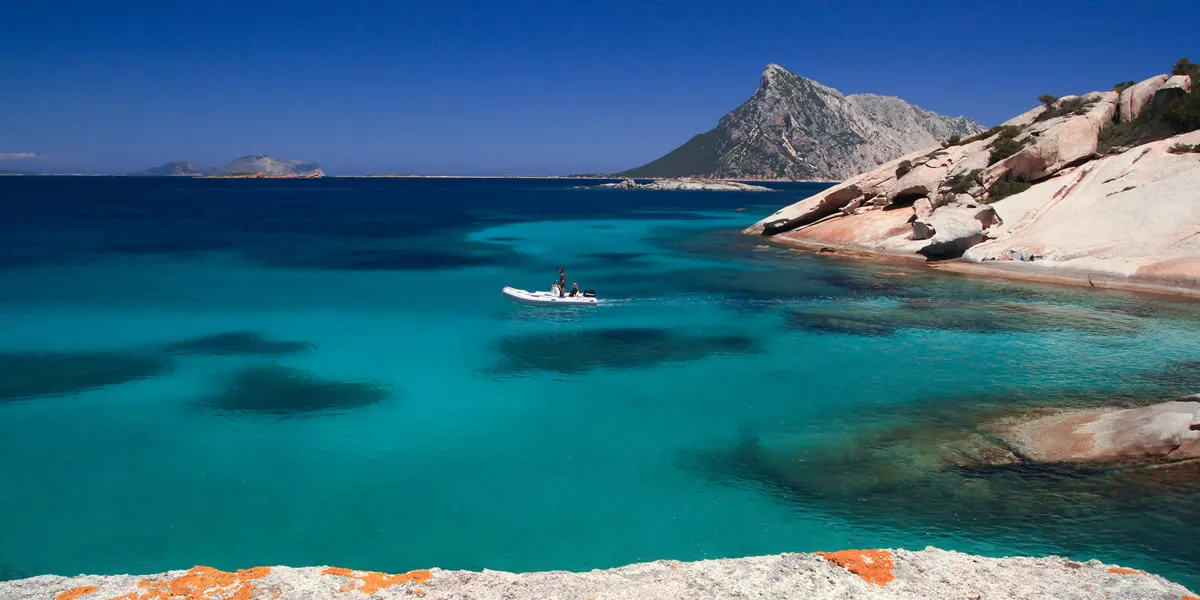 Small boat anchored in a turquoise granite cove with Tavolara island on the horizon