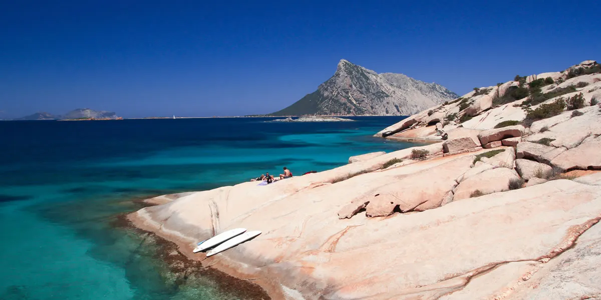 Sunbathers and paddleboards on the flat pink granite slabs of Sassi Piatti, Tavolara island across the deep blue water