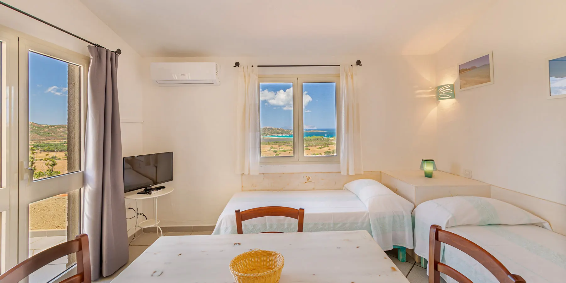 Apartment interior with dining table, beds and a window onto the bay of Porto Taverna