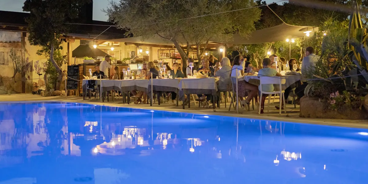 Long dinner table by the pool at night, candlelight reflected in the blue water