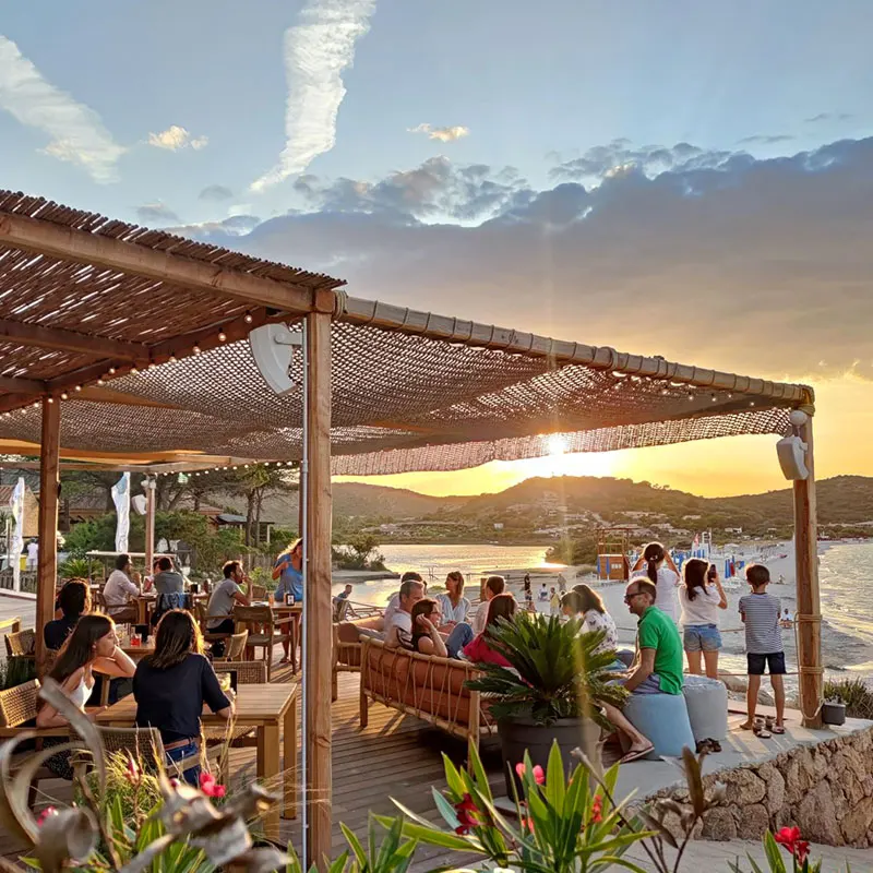 Pergola deck of the Tavernetta beach bar at golden hour, guests looking over Porto Taverna bay